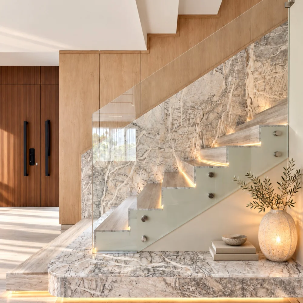 Bottom steps of a staircase cut from veined stone, lit underneath, with a glass balustrade and the walnut entrance doors beyond, in an Al Furjan villa by Tohfa